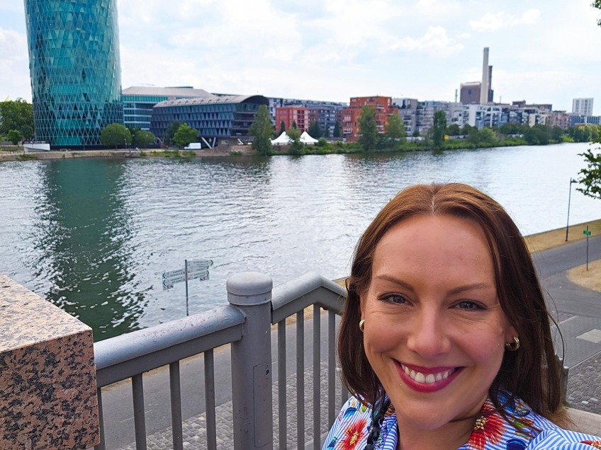Selfie am Mainufer in Frankfurt: Im Vordergrund lächelt eine Frau mit schulterlangen braunen Haaren in die Kamera, im Hintergrund die Skyline mit einem markanten türkis-verglasten Hochhaus und moderne Wohn- und Bürogebäude entlang des Flusses.